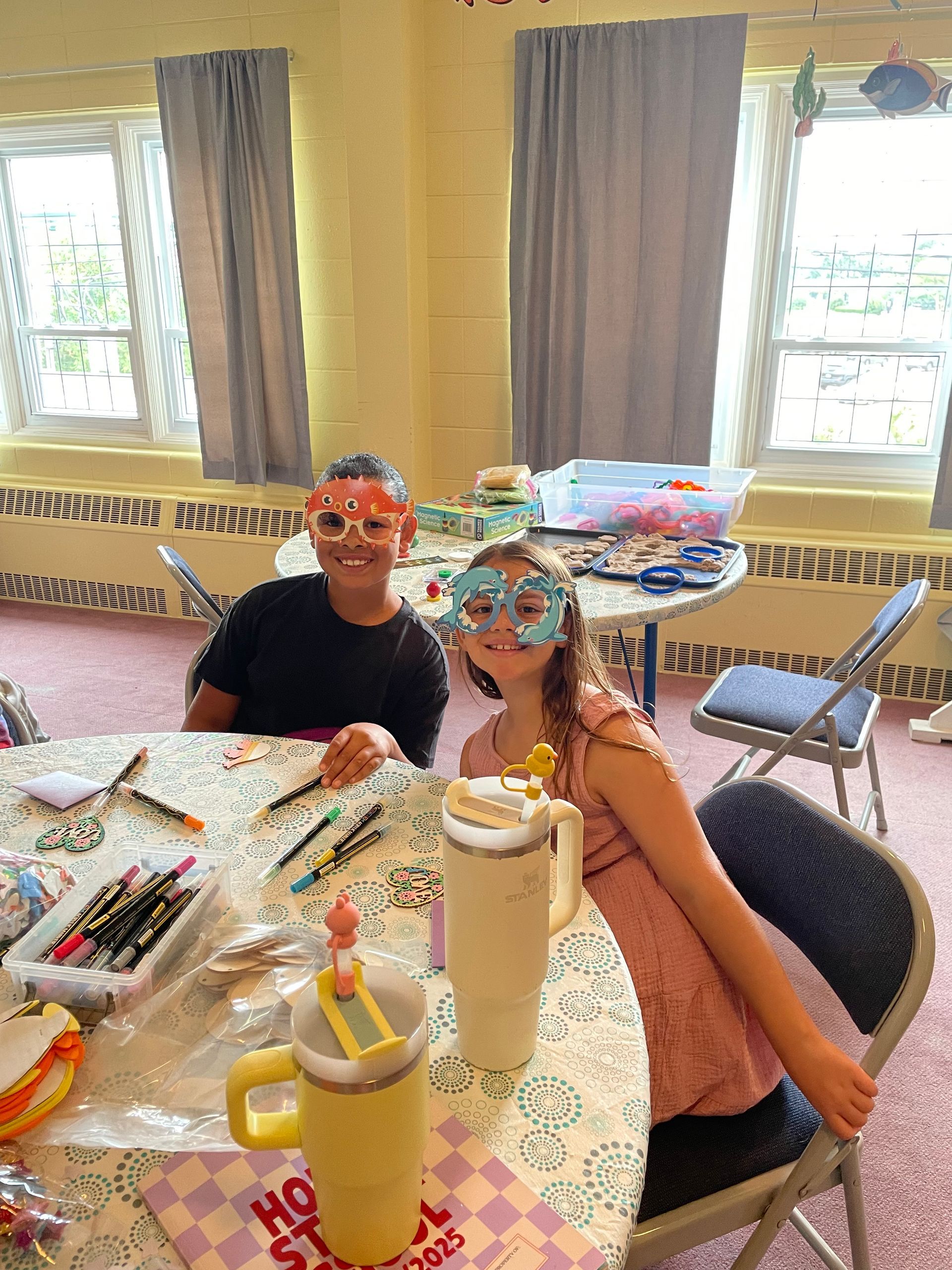 Two people wearing colorful masks sit at a craft table covered with supplies in a bright room.