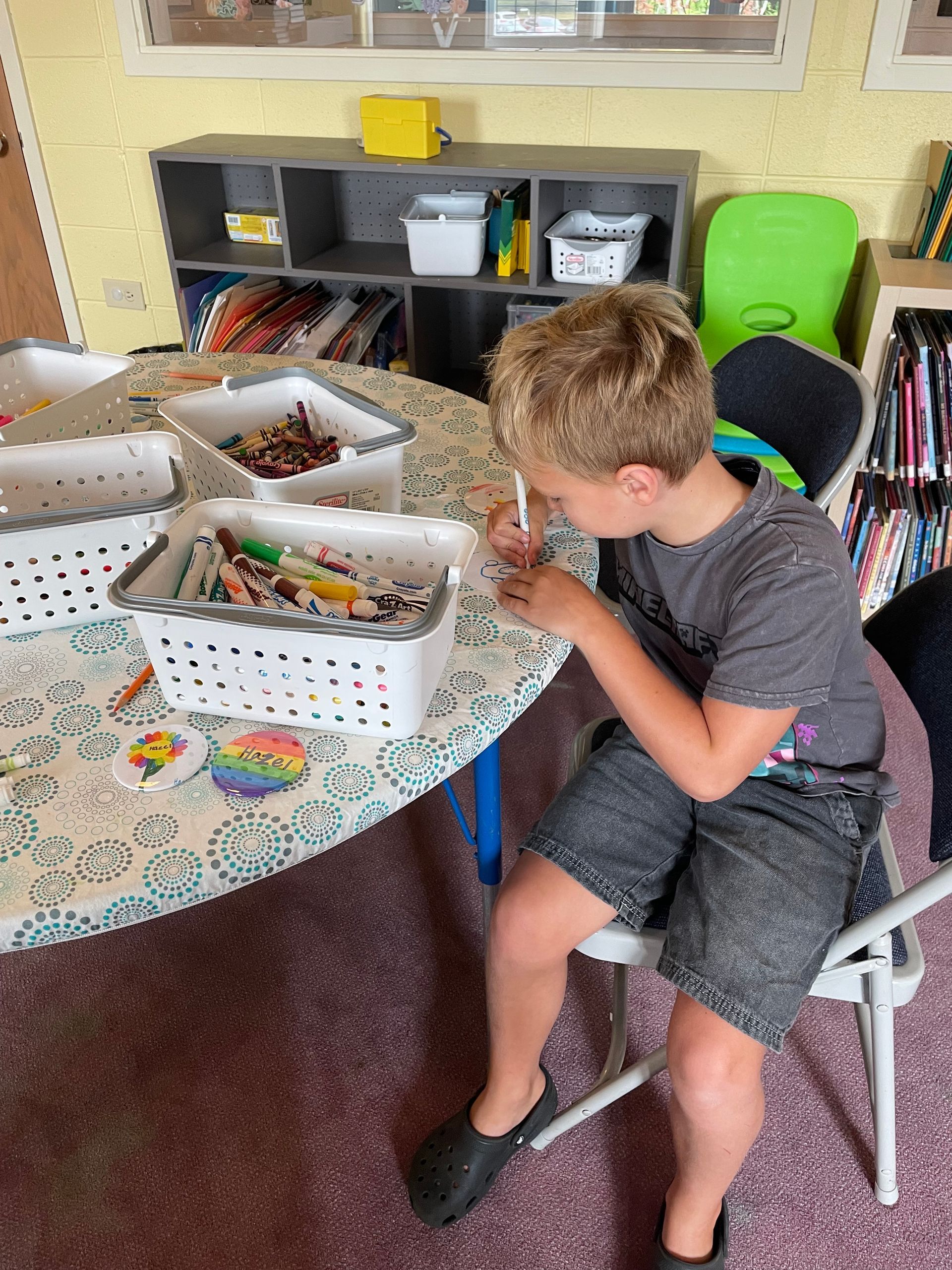 Child sitting at a table, examining craft supplies in white baskets at a play area.