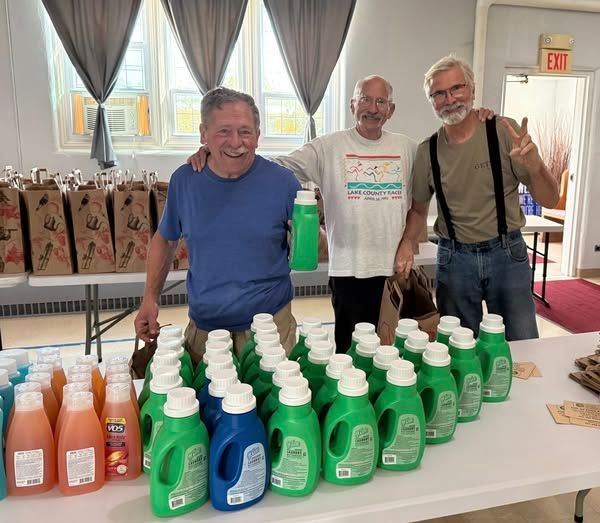 Three men stand behind rows of laundry detergent bottles at a table indoors.