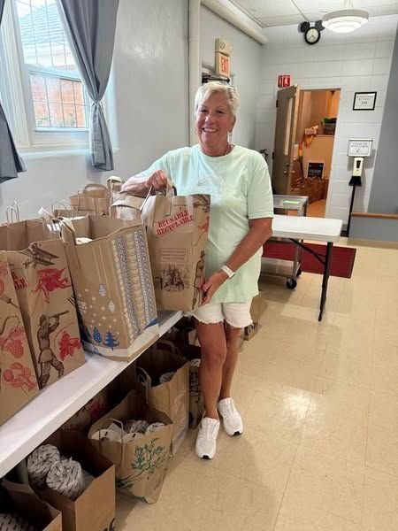 Person smiling in a hallway, standing by a table of gift bags and boxes holding a wrapped package