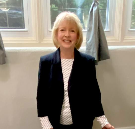 A woman in a suit and striped shirt is standing in front of a window.