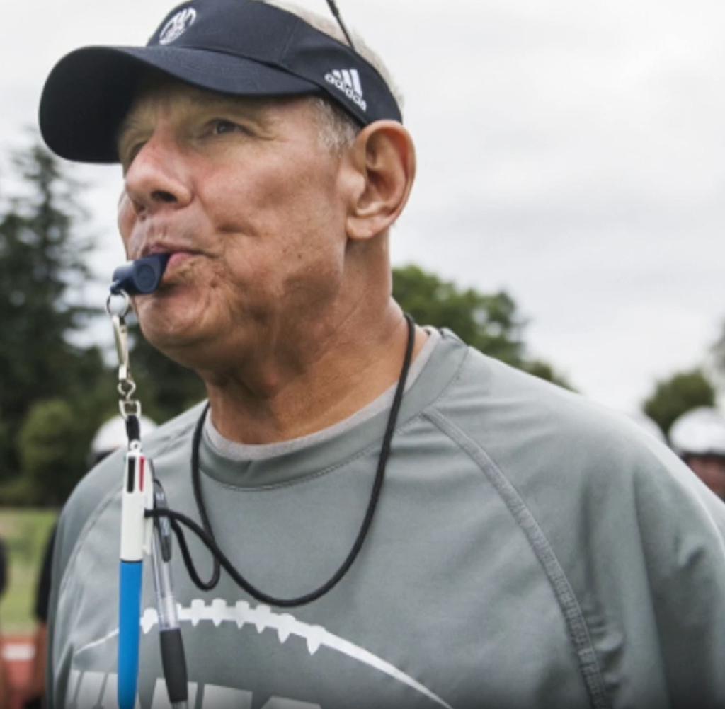 Coach wearing a visor blowing a whistle, in gray shirt, outdoor setting.