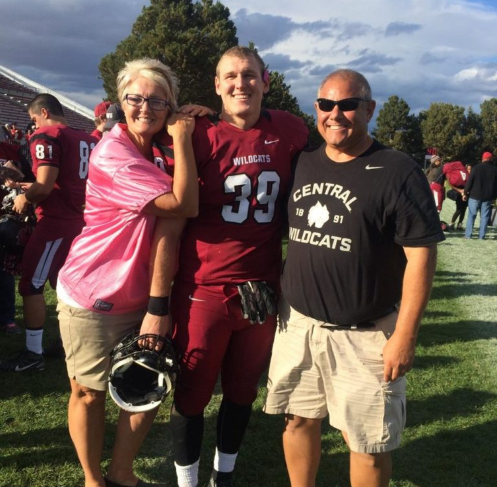 A football player in maroon uniform poses with his parents on a field.