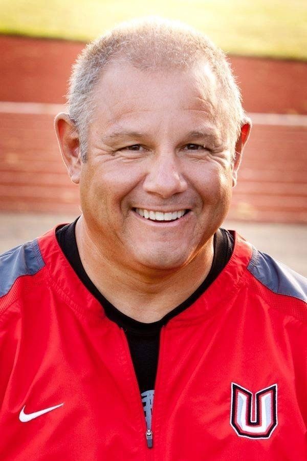 Coach Mark Rego in his red Union High School jacket, smiling at the track.