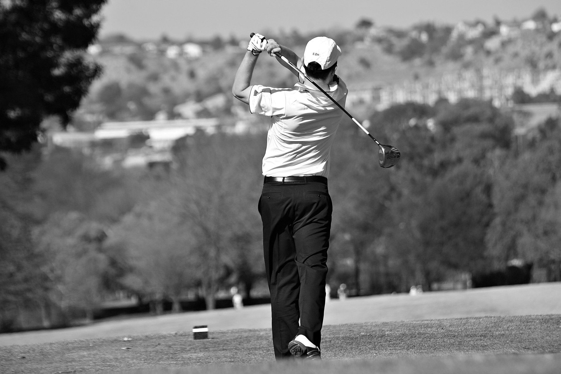 Golfer swinging a club on a course, trees, hills and sky in background. Black and white.