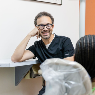 Dentist in black scrubs smiles at a patient in a dental office.