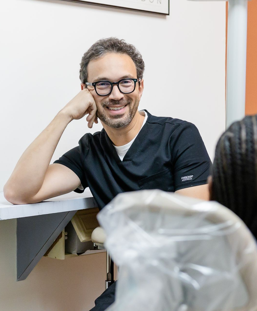 Dentist in black scrubs, glasses, smiling, leaning on dental chair, patient partially visible.