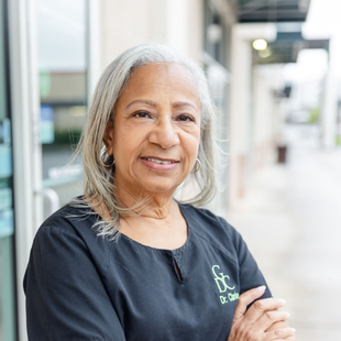 Woman with gray hair smiles, arms crossed, outside a building with glass doors, wearing a black shirt with a logo.