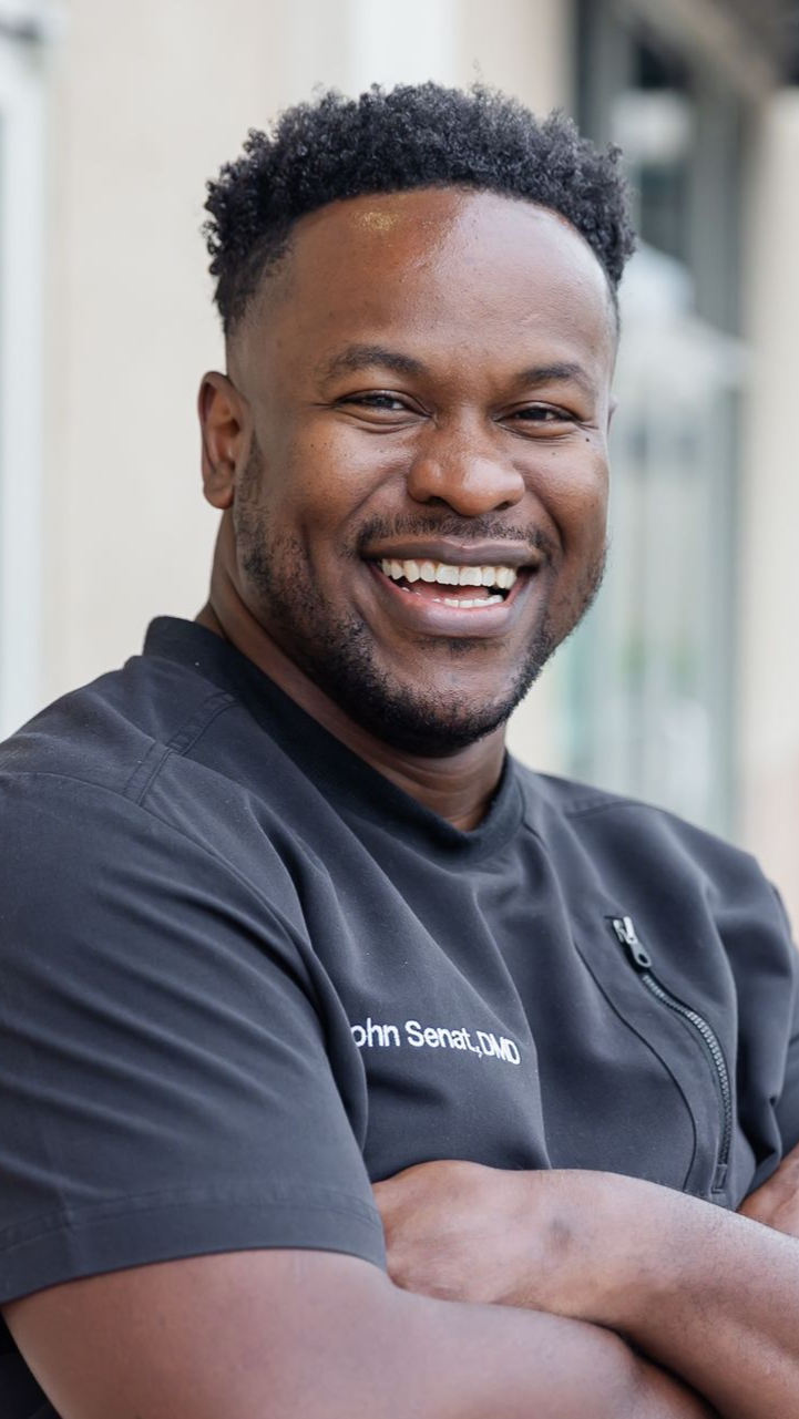 Man smiling with arms crossed, wearing a black shirt with a zipper, in a bright outdoor setting.