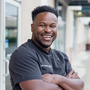 Man in black scrubs smiles with arms crossed, standing outside a building.