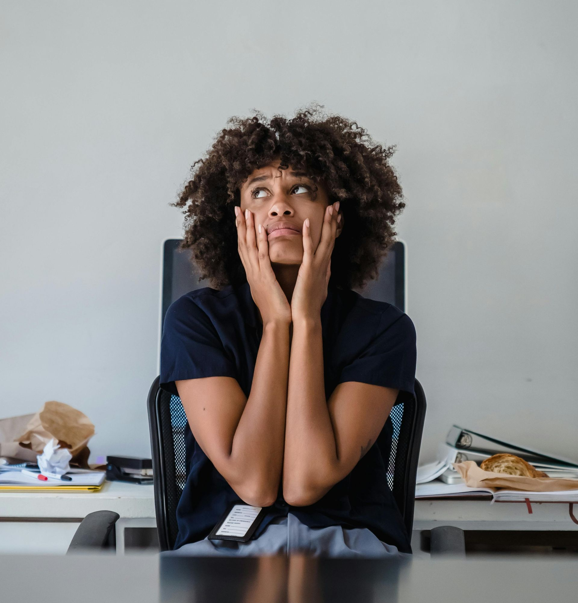 Woman with curly hair, hands on cheeks, looking upward in a cluttered office environment.