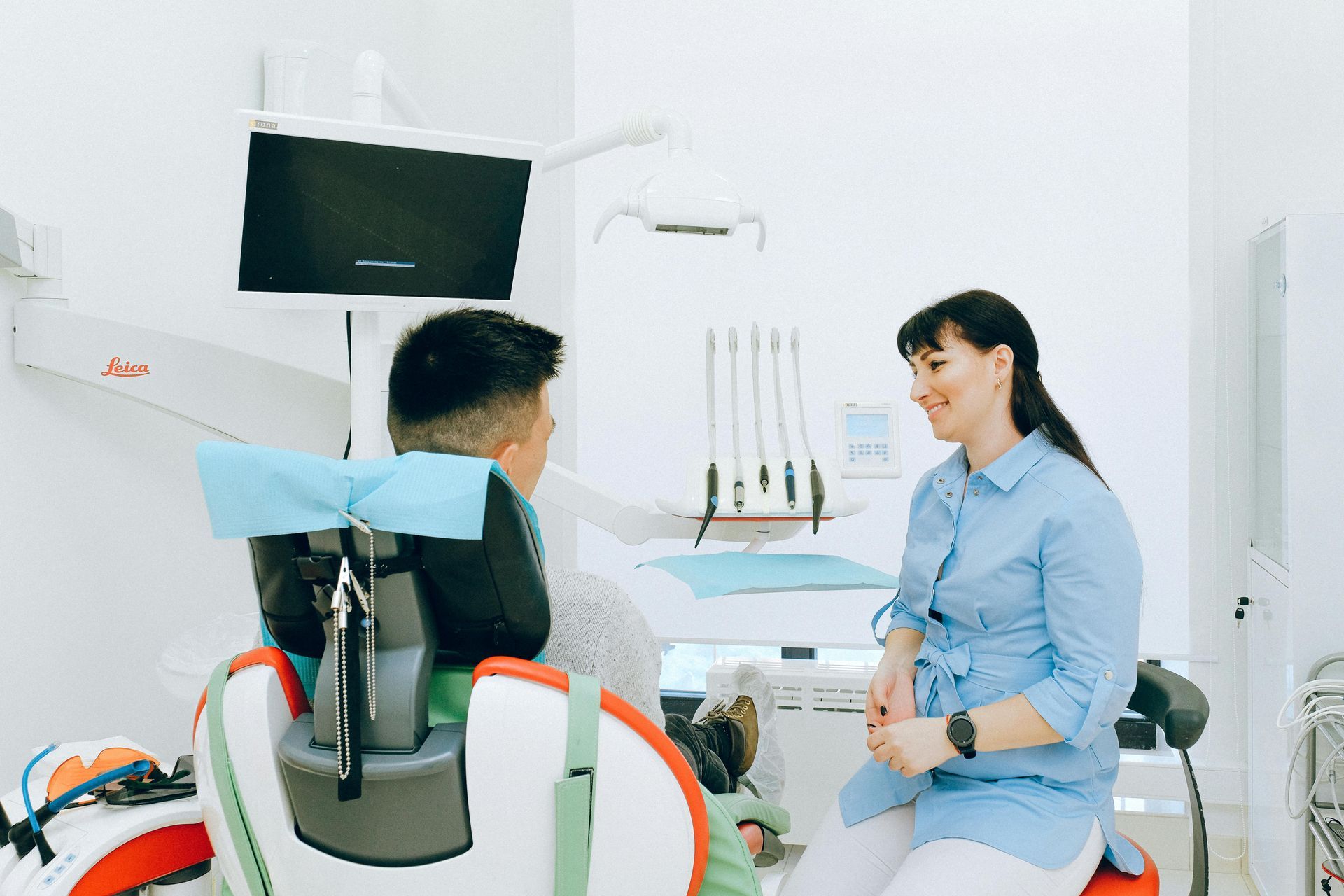 Dentist in blue shirt talking to patient seated in a dental chair in a bright, clean dental office.