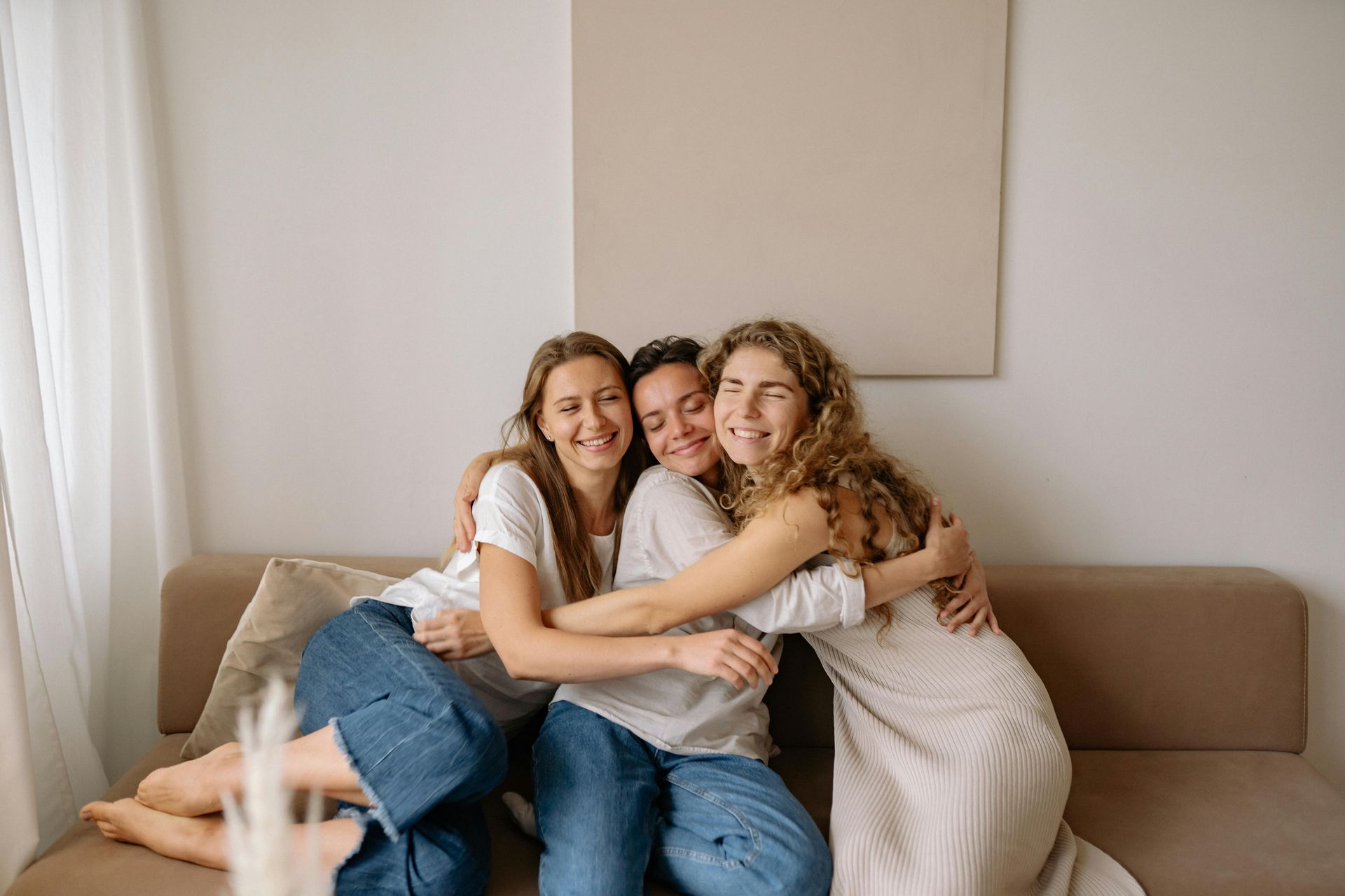 Three friends hugging on a beige sofa. White-walled room, light-colored clothing, happy expressions.