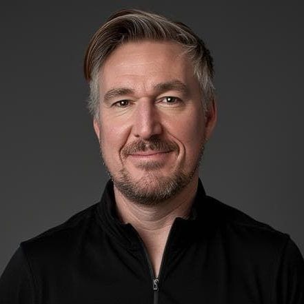 Man with short, graying hair and beard smiles in a black shirt against a gray background.