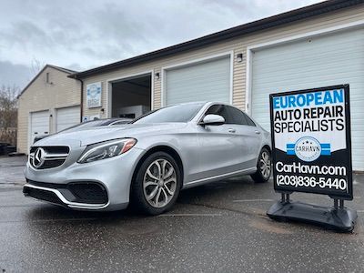 A silver mercedes benz is parked in front of a garage next to a sign for european auto repair specialists.