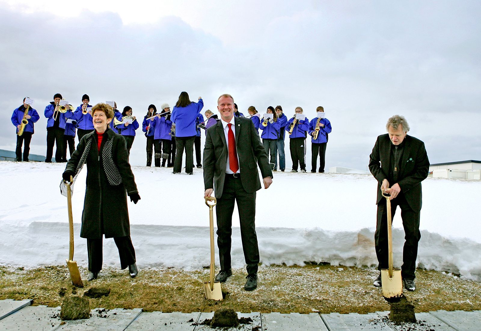 Three people with shovels at the groundbreaking ceremony for Hljómahöll, with a brass band performing in the background.