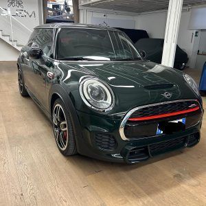 Una Mini John Cooper Works hatchback verde scuro parcheggiata all'interno di uno showroom di una concessionaria illuminato a giorno.