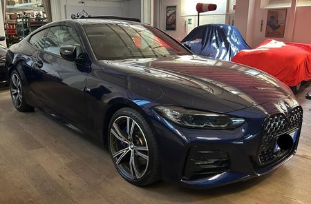 Una BMW Serie 4 coupé blu scuro parcheggiata all'interno di un garage, accanto a veicoli coperti da teli protettivi blu e rossi.