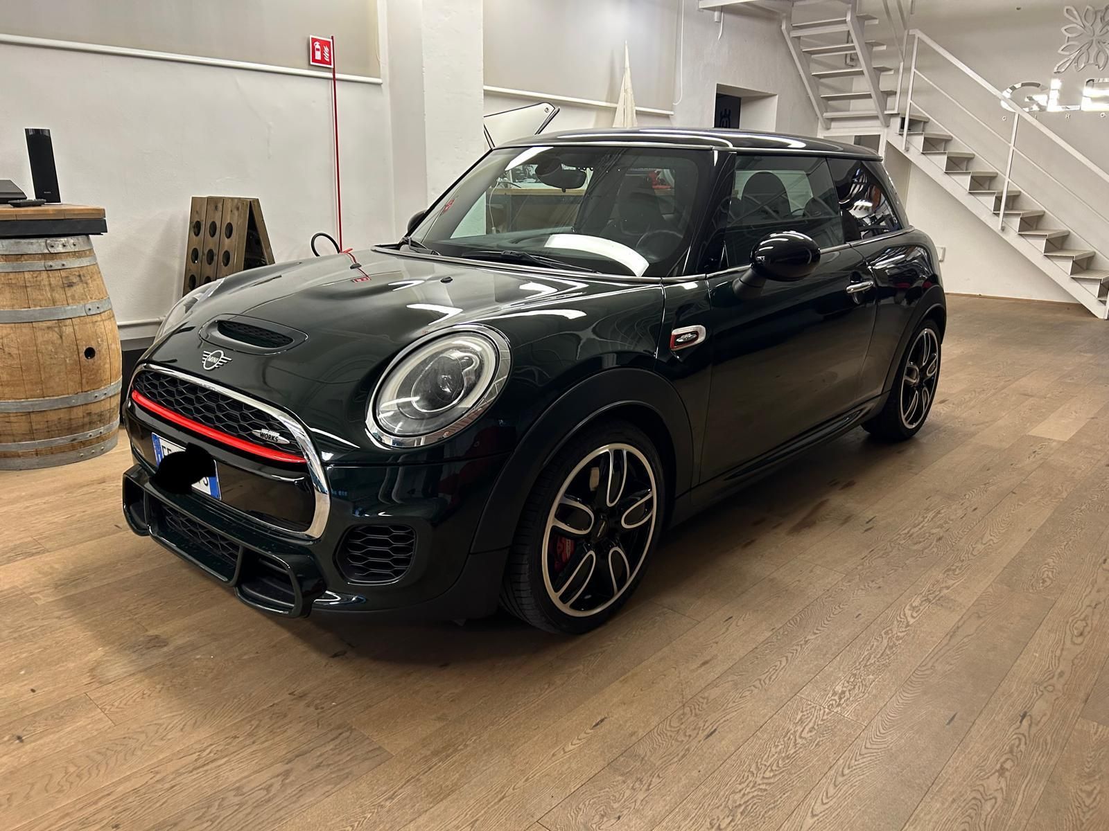 Una Mini Cooper verde scuro con finiture rosse sulla griglia anteriore, parcheggiata in un luminoso e moderno showroom al coperto con pavimento chiaro.