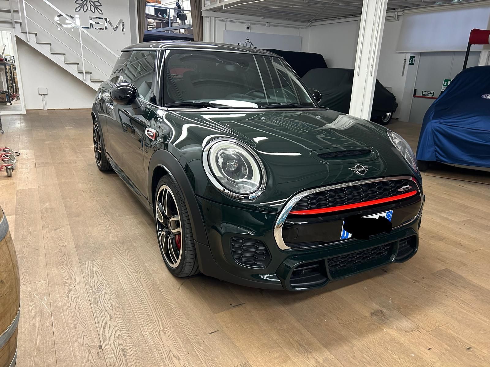Una MINI Cooper verde scuro parcheggiata in un garage con pavimento in legno, con una vista parziale di una scala e un'auto coperta.