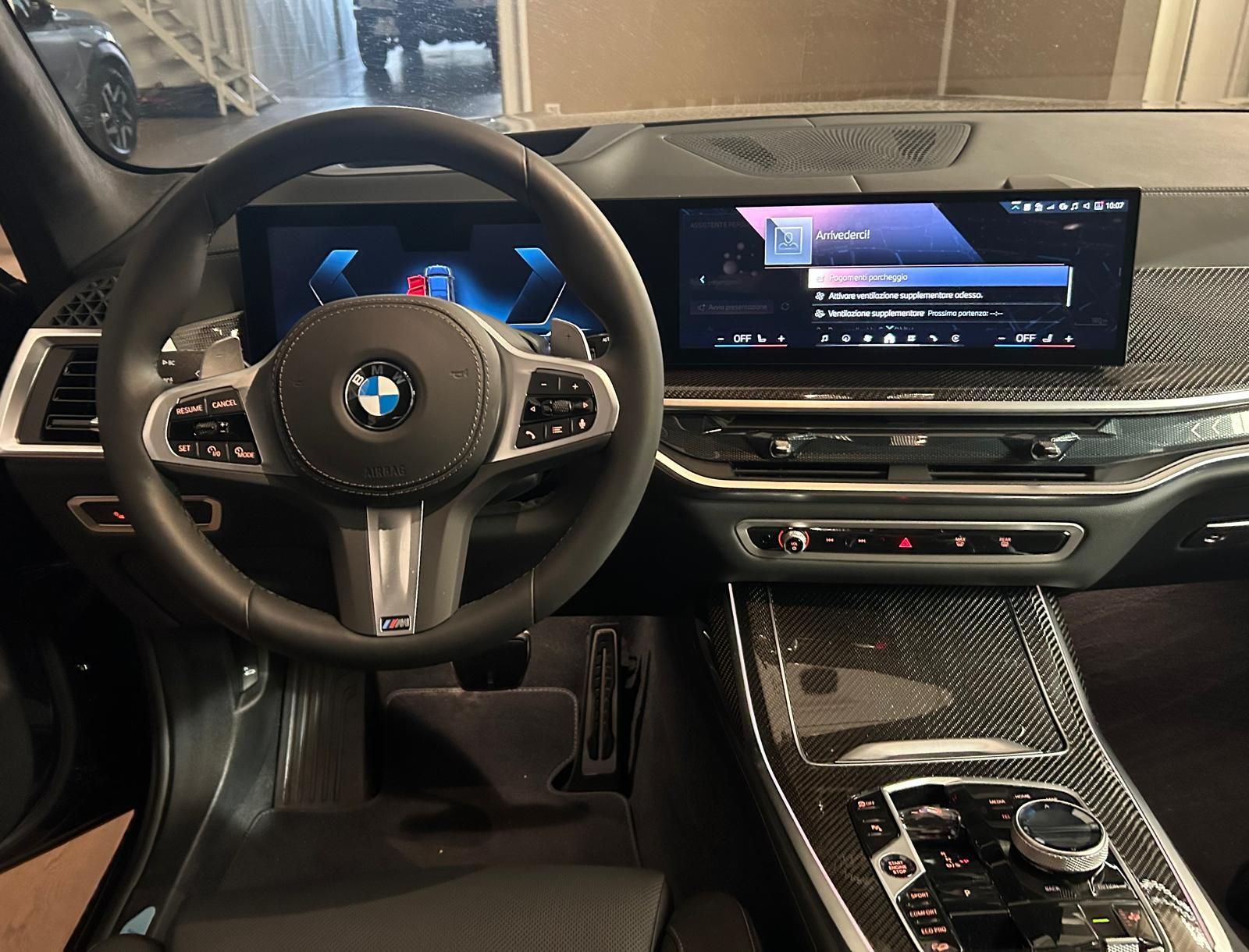 Vista dal posto di guida di un moderno abitacolo BMW, con cruscotto digitale, volante in pelle e finiture in fibra di carbonio.