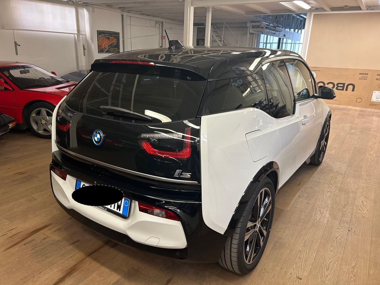 Una BMW i3 bianca e nera parcheggiata all'interno di un edificio, su un pavimento in legno, con un'auto sportiva rossa visibile sullo sfondo.