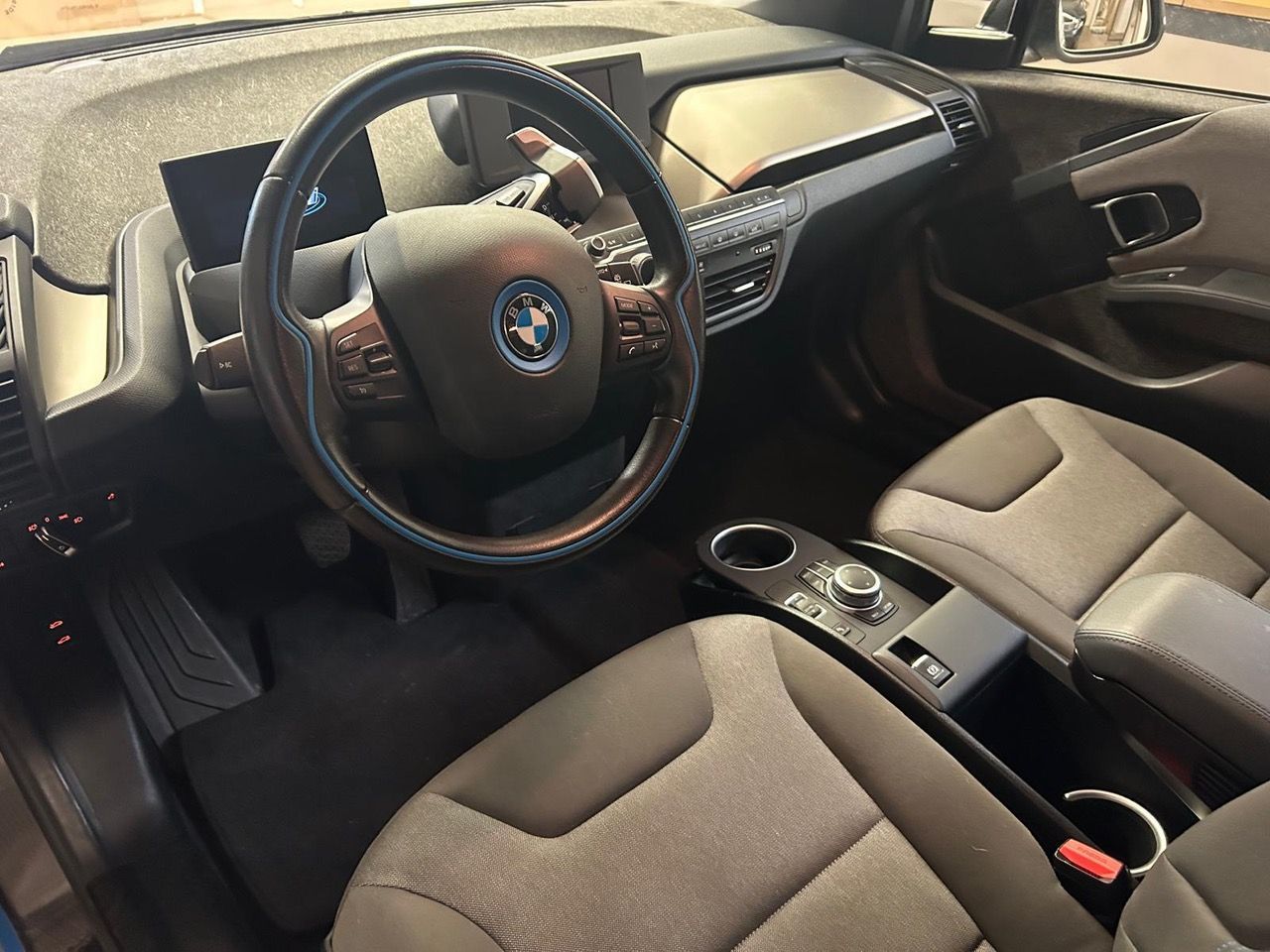 Vista interna di una BMW i3 con volante scuro con logo blu, sedili in tessuto grigio e cruscotto moderno.