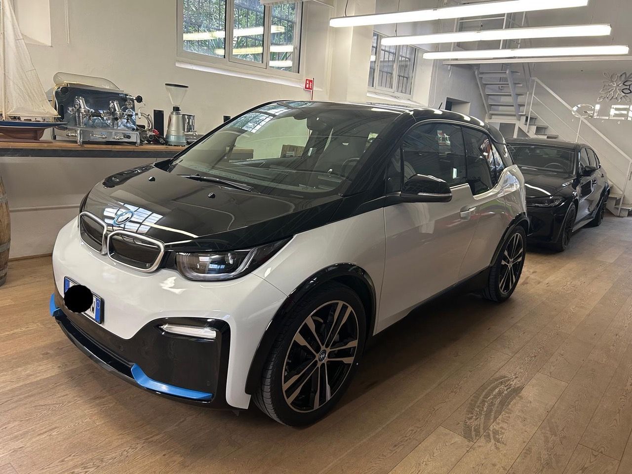 Una BMW i3 bianca con tetto, cofano e finiture nere, parcheggiata all'interno di uno showroom moderno con pavimenti in legno e illuminazione brillante.