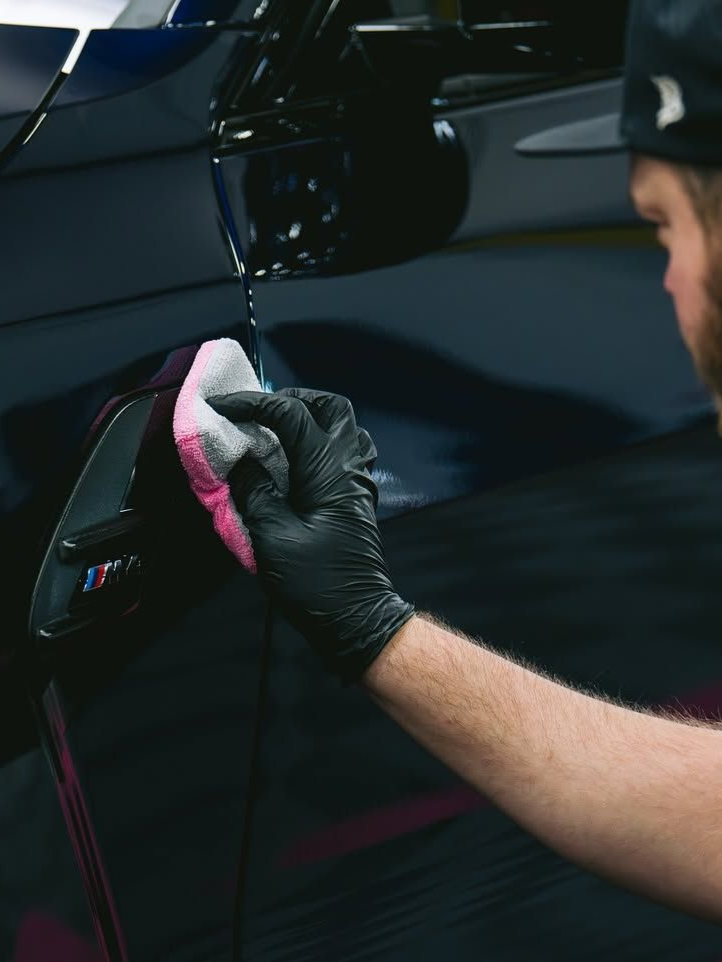 Gloved detailer polishing a black car door with a pink microfiber cloth