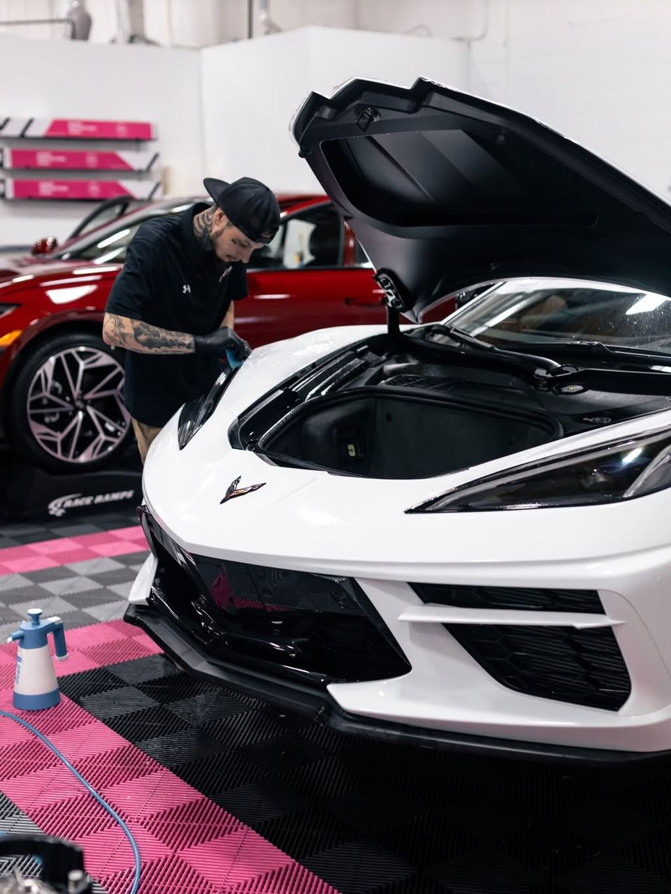 Person working on a white sports car in a garage, with a red car and cleaning spray nearby