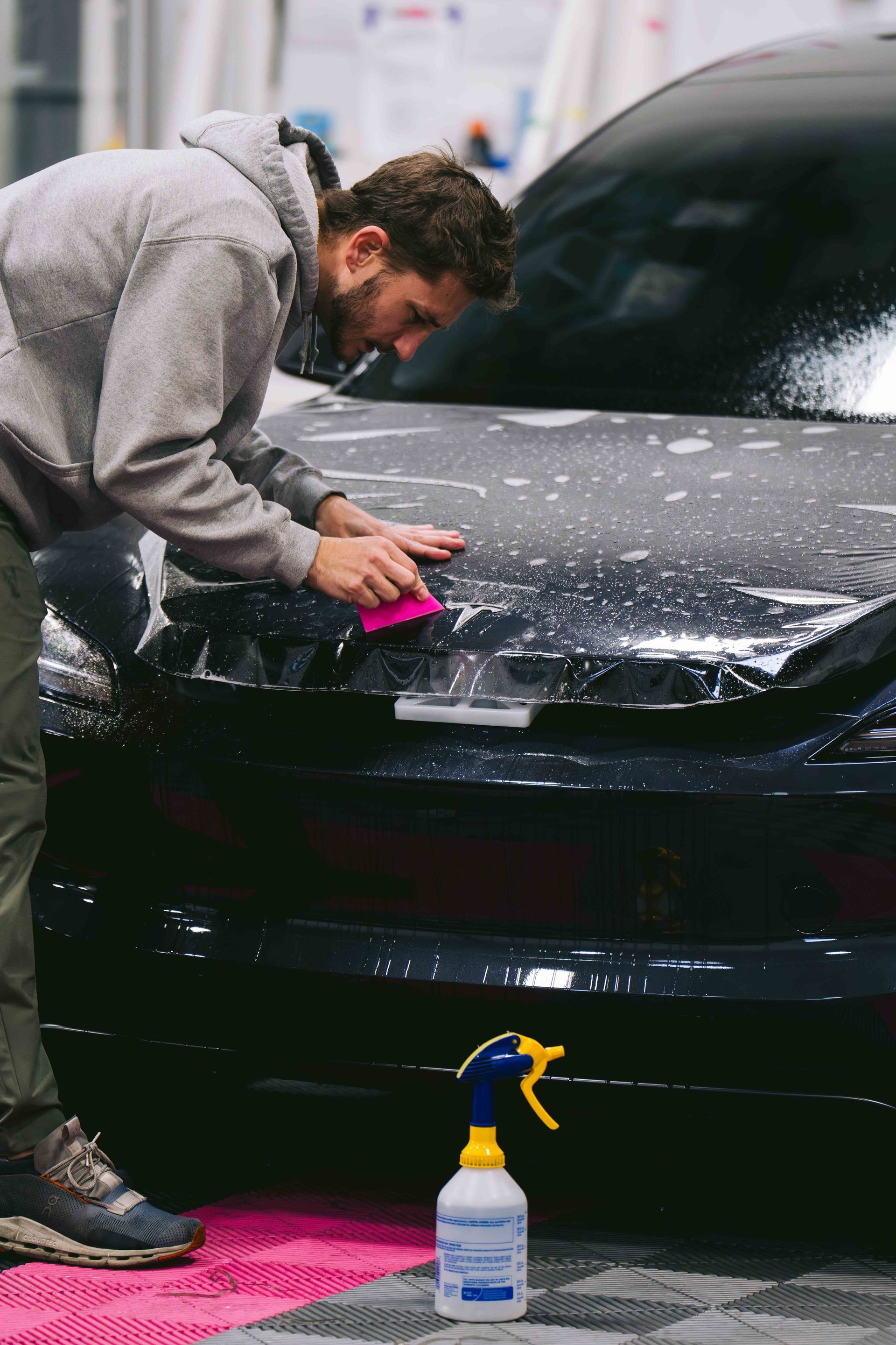 Person hand-washing a black car with a pink sponge and spray bottle in a garage.