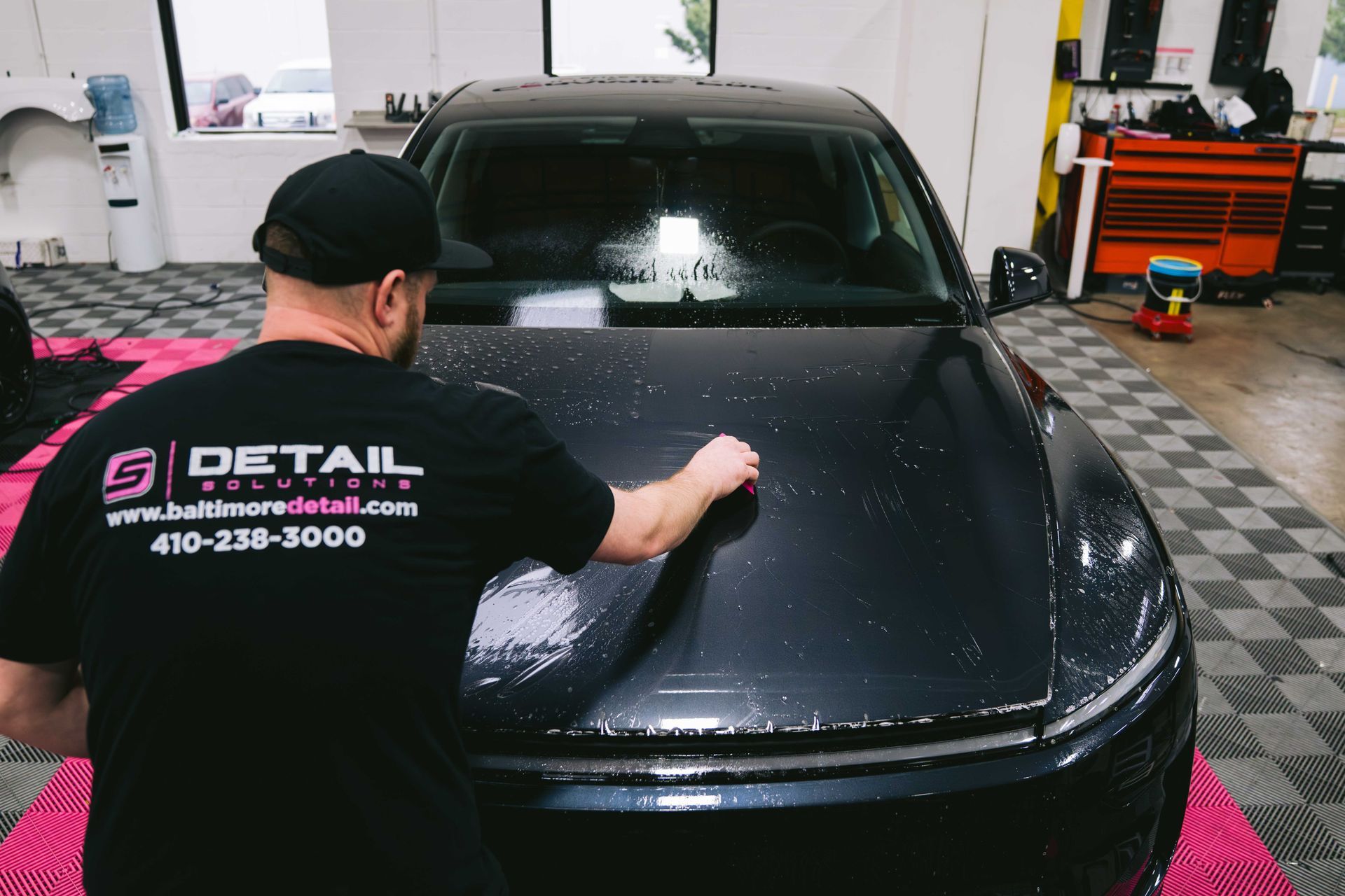 Detail worker polishing a black car hood in an auto shop with checkered floor mats