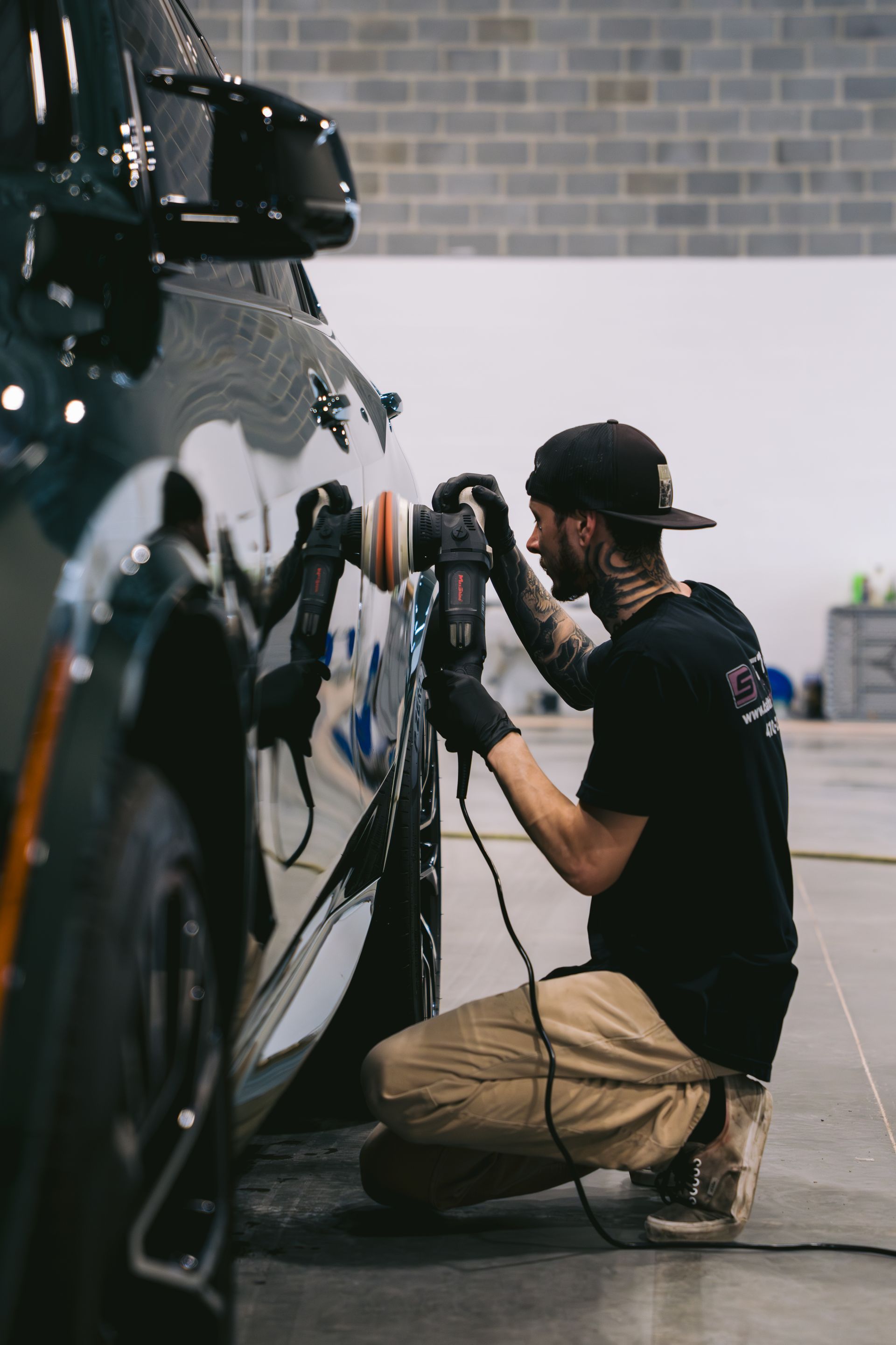 Person kneels beside a black car, polishing the door in a garage with a brick wall backdrop.
