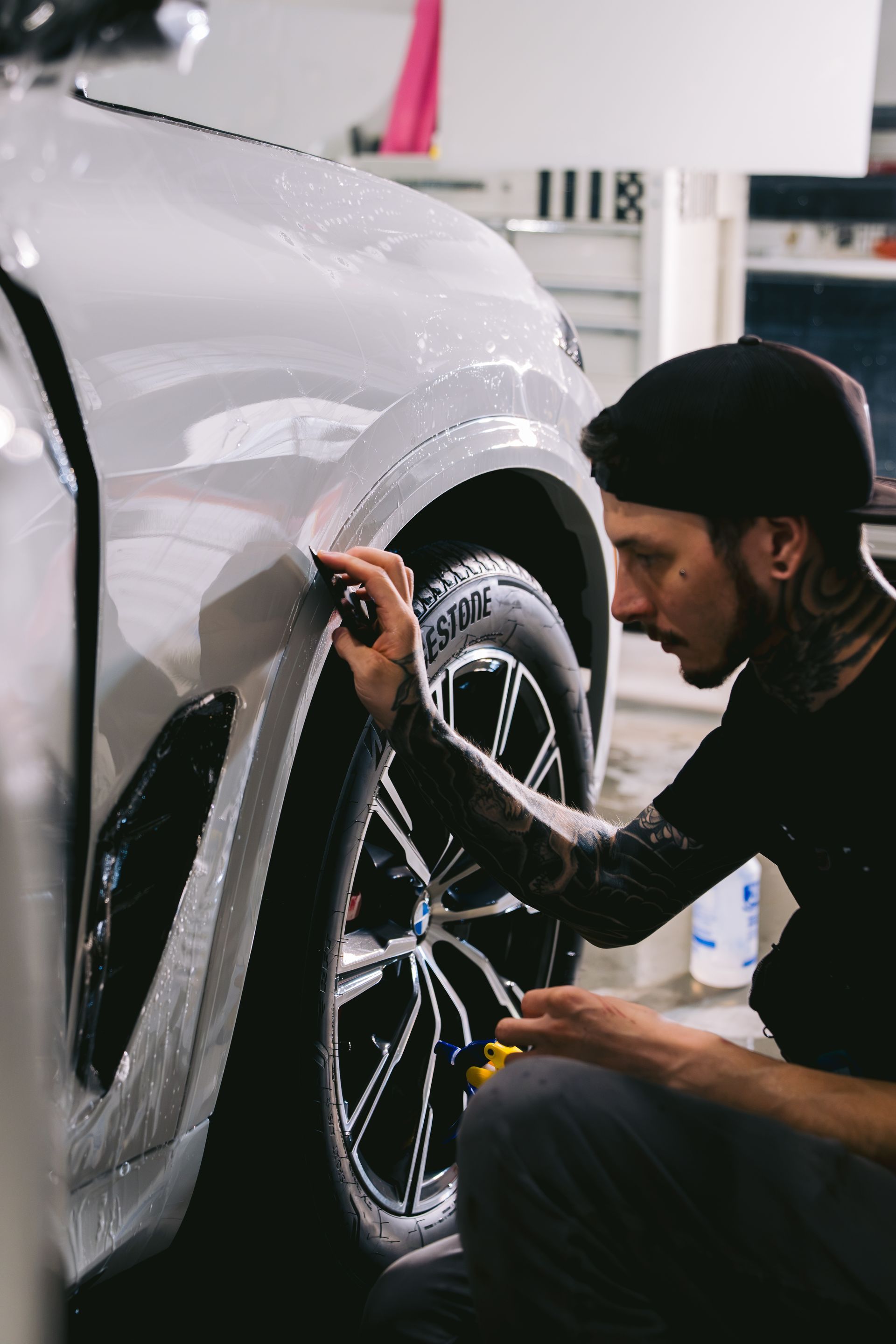 Mechanic polishing a car headlight in a garage, wearing a black cap and shirt.