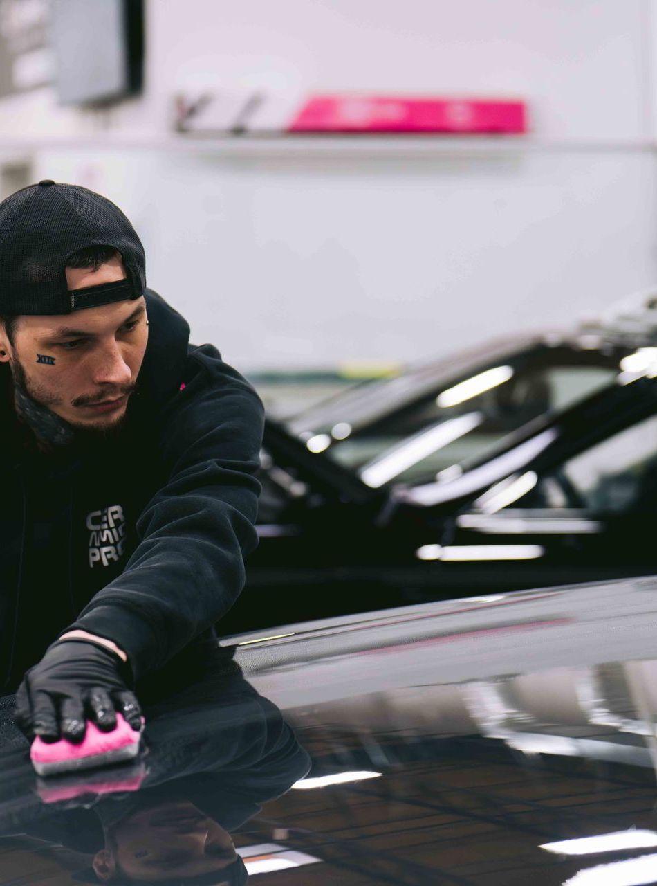 Person applying protective film to a black car's hood. Pink tool, bubbles visible, indoors.