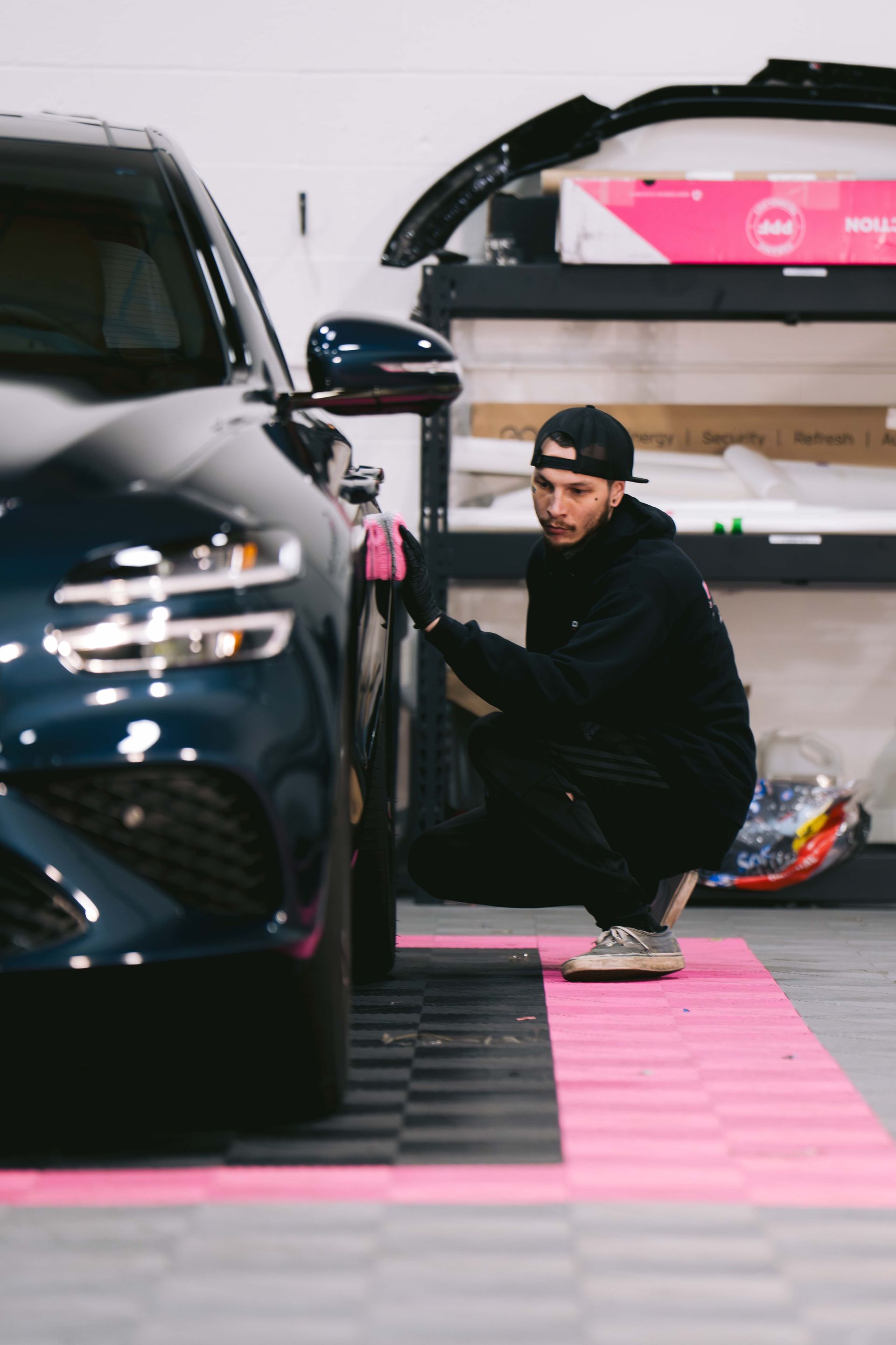 A person in a black hoodie and cap crouches beside a dark blue car in a garage, cleaning the exterior with a pink cloth.