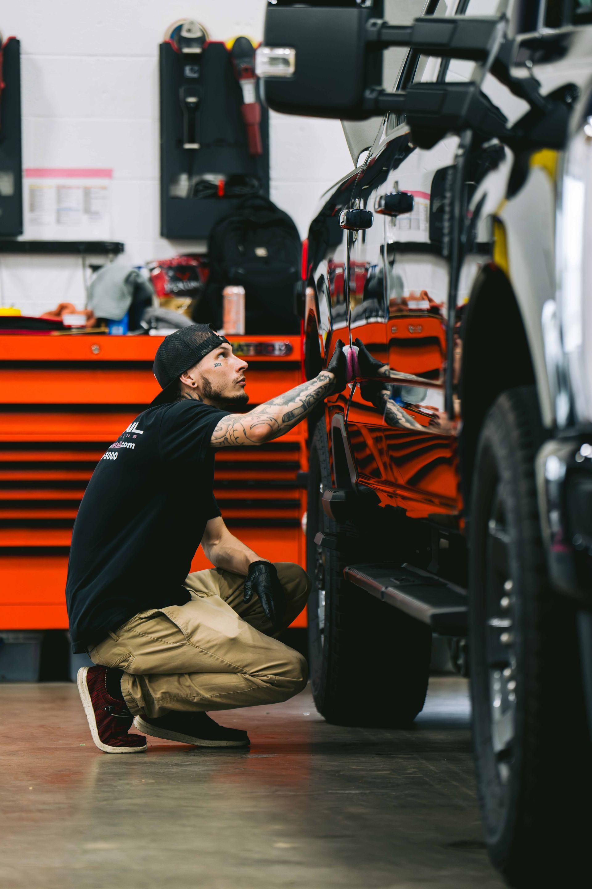 A person wearing a black shirt and cap crouches beside a truck, applying a buffing tool to its side in a workshop.