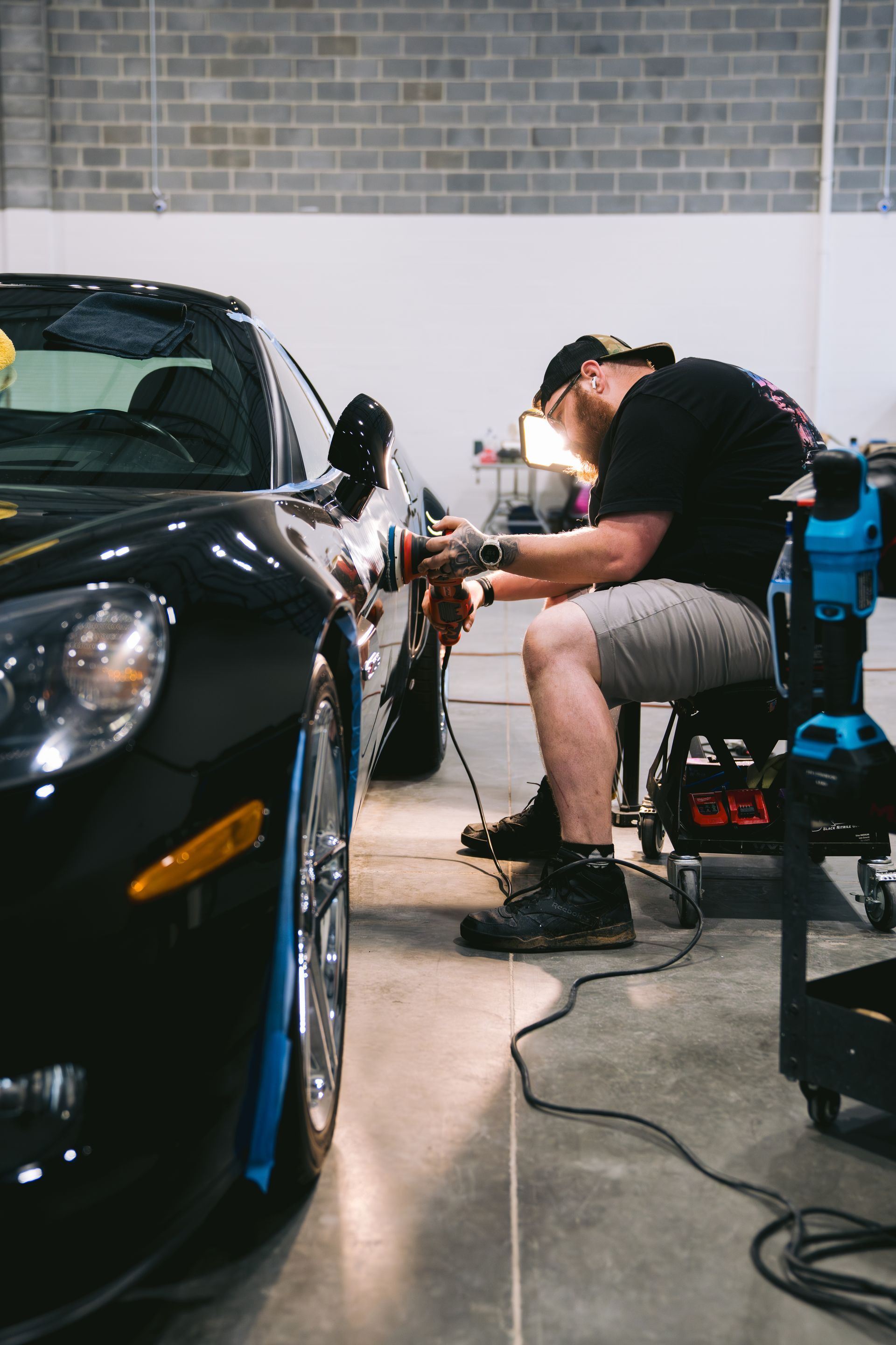 Technician polishing a black car hood in a workshop, with bright lights in the background