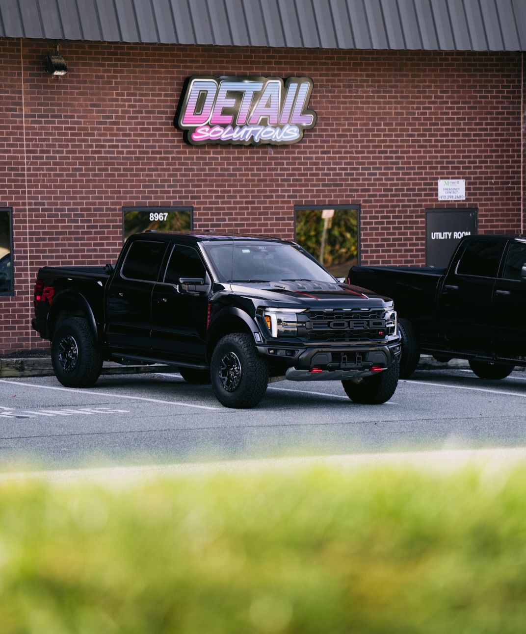 Black pickup truck parked outside a brick detail shop with a neon sign.