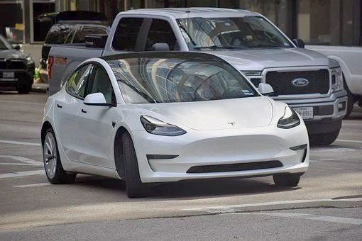 a white tesla model 3 is driving down a city street next to a ford f150 truck .