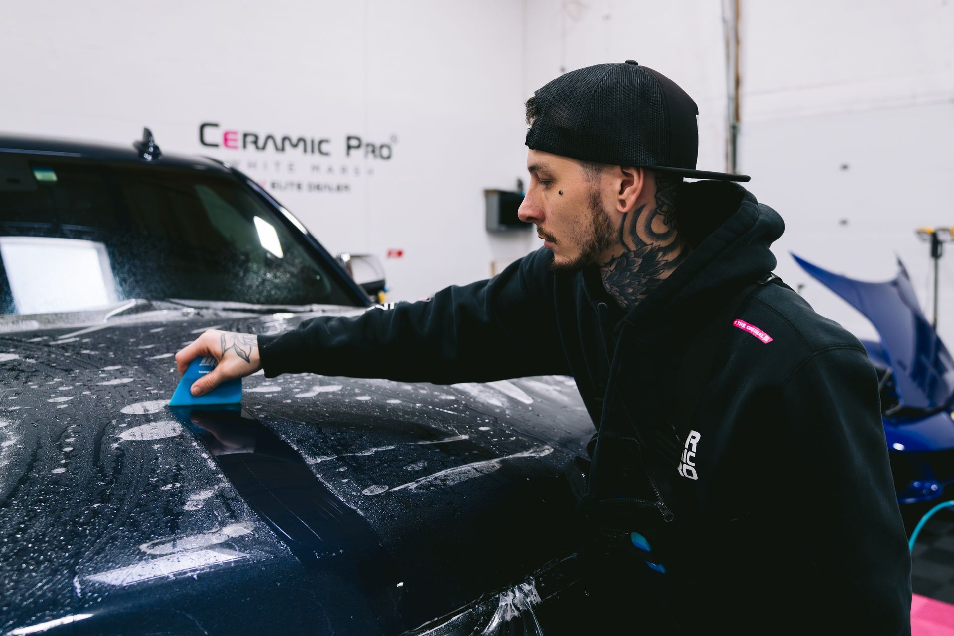 Person in a black hoodie and cap cleaning a dark car hood in a detailing shop