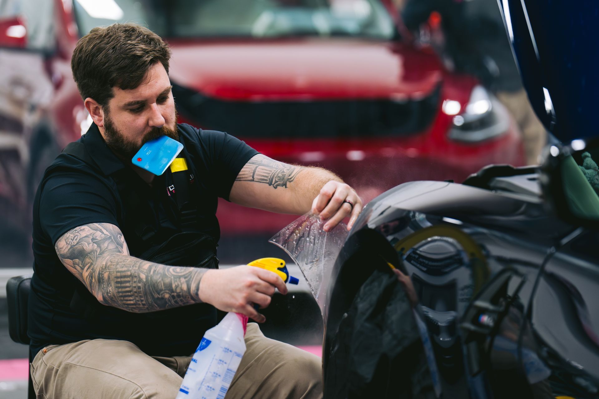 Man applying film to a black car fender with spray bottle. Workshop setting with red car in background.