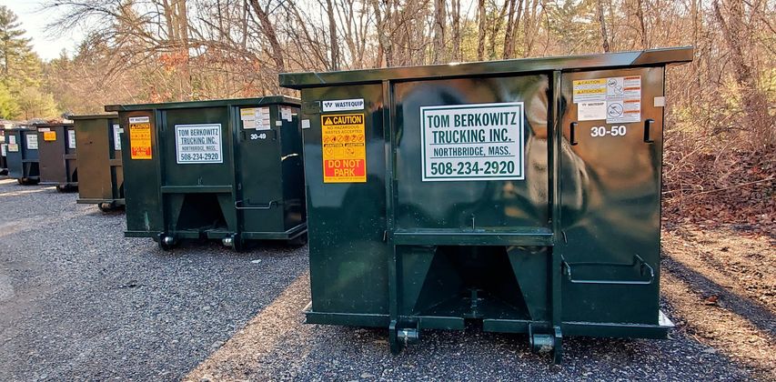 Several dark green dumpsters lined up on a gravel lot near trees.