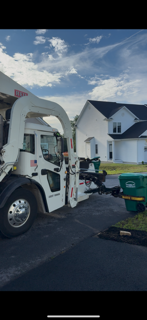 A white garbage truck is parked on the street next to green trash bins under a blue sky.
