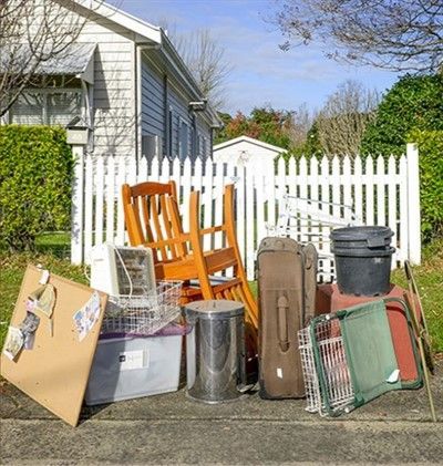 Pile of discarded furniture and household items on a curb in front of a white picket fence and house.