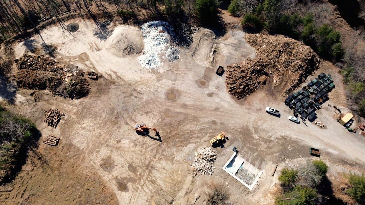 Aerial view of a clearing with construction equipment, piles of wood, and a concrete foundation.