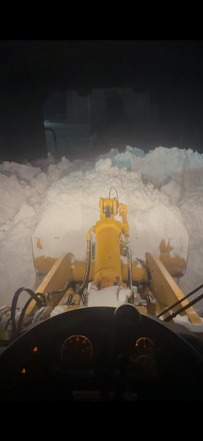 Snowplow pushing through deep snow at night, viewed from the cab with headlights illuminating the snowbank.