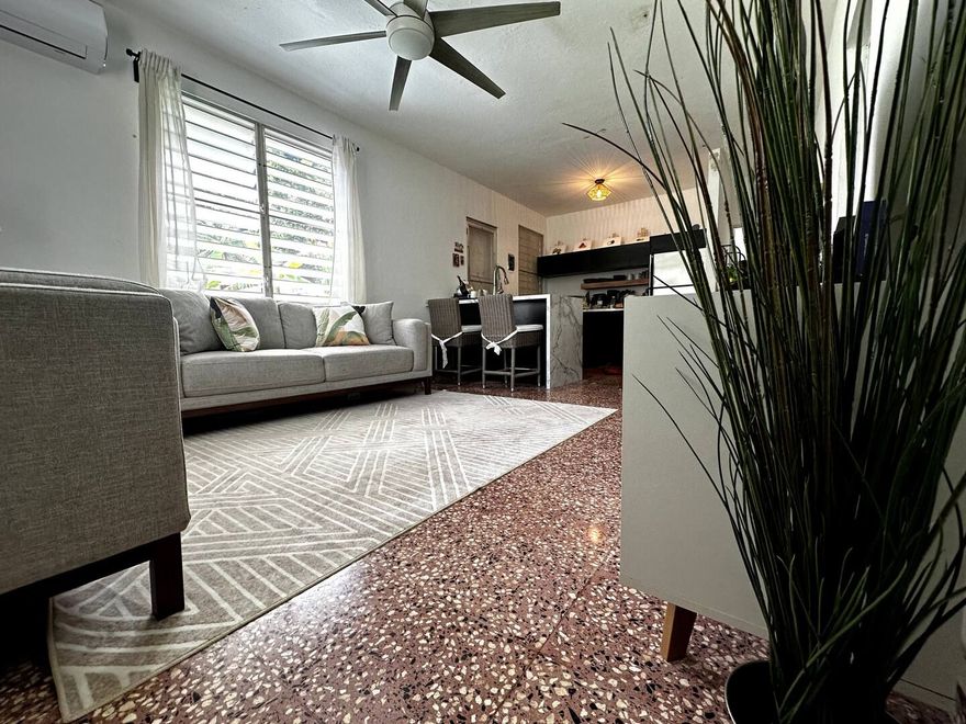 A living room with a grey sofa on a patterned rug, terrazzo flooring, and a dining area with bar stools in the back.
