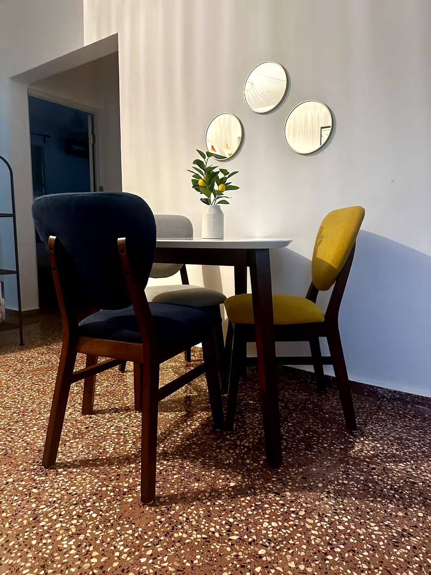 A small dining table with one navy and one mustard chair on a speckled floor, with three round mirrors on a white wall.