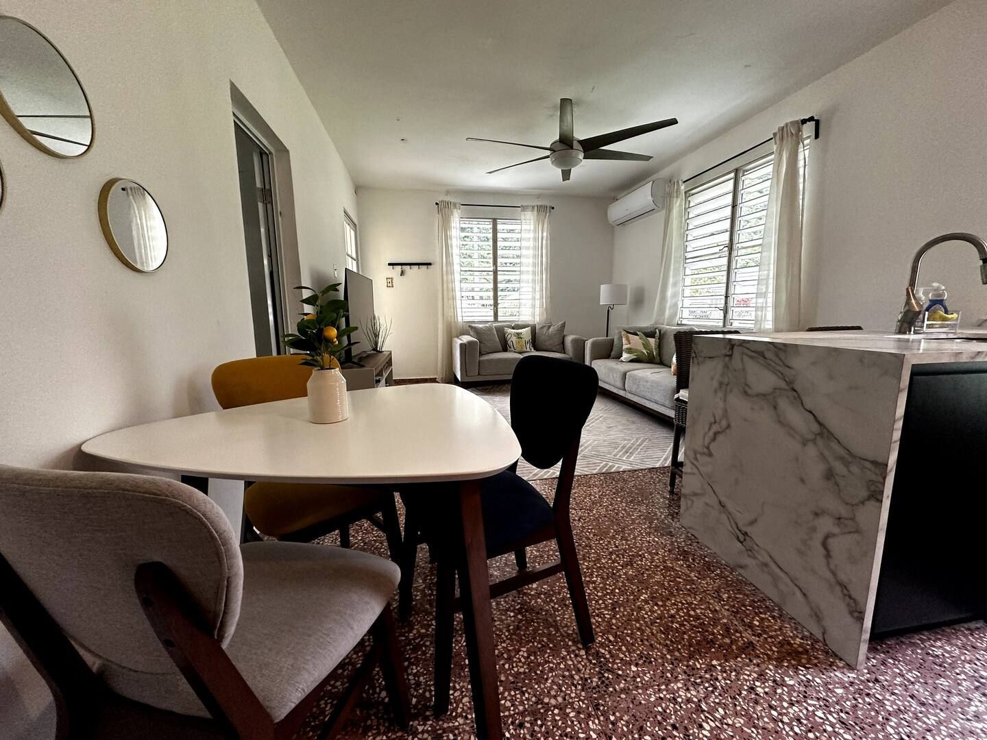 A brightly lit dining and living area with a white table, chairs, marble-patterned island, and terrazzo flooring.